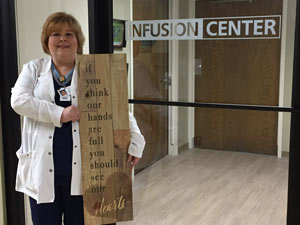 This is a picture of Jessica Eldridge holding a sign that says if you think our hands are full you should see our heart while standing at the entrance of the infusion center